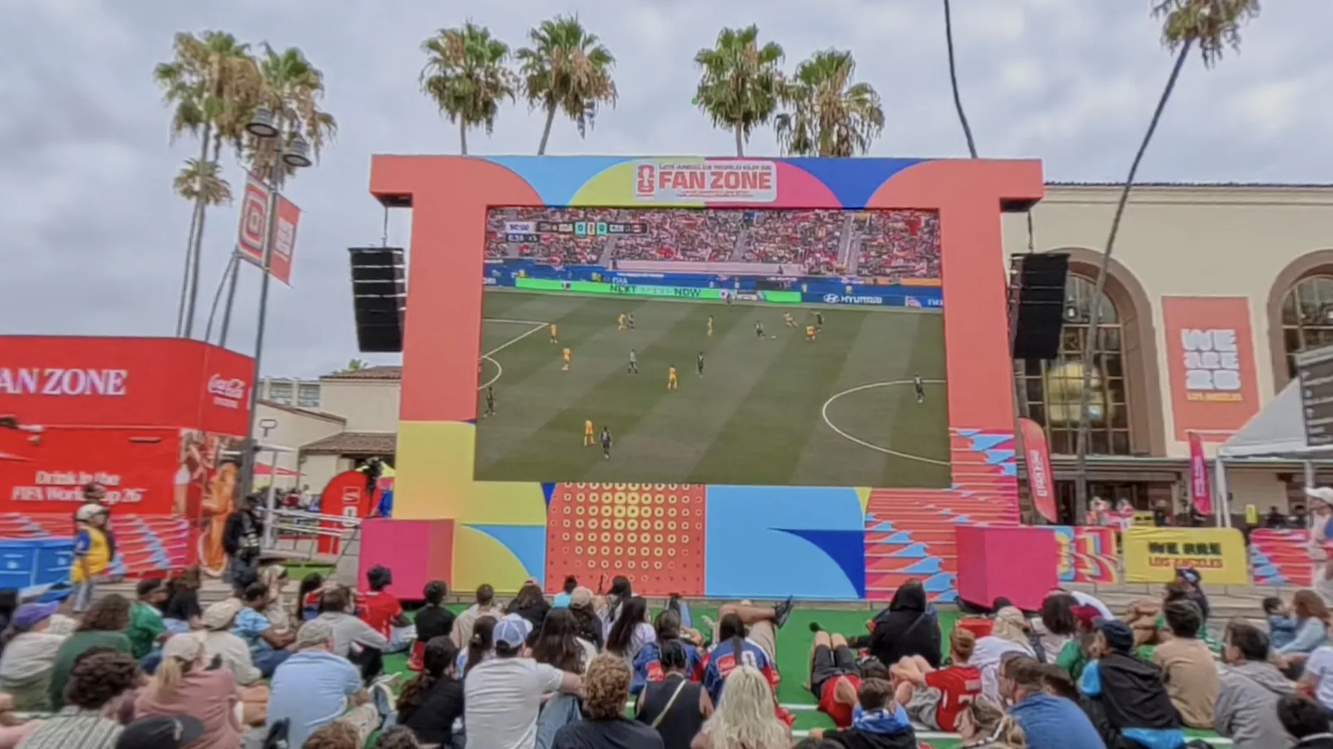 The athlete's view inside the headset of a World Cup fan zone, with a crowd seated in front of a large screen showing a live match